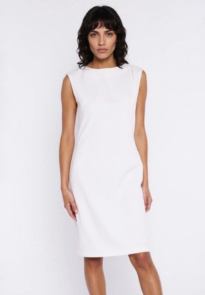 Kobieta z krótkimi falistymi ciemnymi włosami, ubrana w białą, bezrękawową sukienkę do kolan, stojąca na jednolitym białym tle.