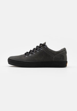 Niskie ciemnoszare zamszowe sneakersy z czarną gumową podeszwą, czarnymi sznurowadłami przewleczonymi przez metalowe oczka i subtelnym tłoczonym logo na pięcie.