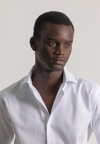 Camisa blanca de botones con cuello puntiagudo, que presenta una textura suave y líneas nítidas, llevada por una persona con cabello corto y oscuro.