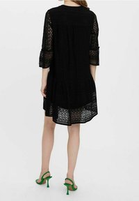 Femme debout, portant une robe en dentelle noire avec des manches trois-quarts et des sandales à talons verts sur un fond blanc uni.