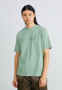 Ανοιχτό πράσινο βαμβακερό T-shirt με ανάγλυφο λογότυπο "Timberland" και έμβλημα δέντρου μπροστά; στρογγυλή λαιμόκοψη και κοντά μανίκια.