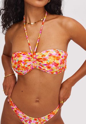 Femme portant un bikini bandeau floral coloré avec un tour de cou, des bijoux en or, debout les mains sur les hanches devant un fond uni.