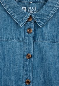 Denim srajca v svetlo modri barvi s koničastim ovratnikom, opremljena s petimi rjavimi gumbi in enostavno, teksturirano blago. Blagovna znamka vidna znotraj ovratnika.