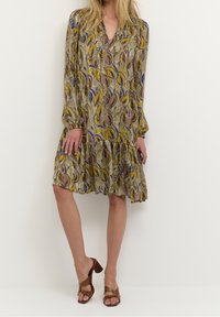 Trykt kjole med lange ærmer, v-udskæring og lagdelt kant. Har et bladmønster i gul, blå og pink på en brun baggrund.