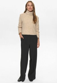 Ženska stoji oblečena v bež pulover s turtleneck izrezom, črne široke hlače in črne čevlje, z eno roko v žepu, na nevtralni ozadju.