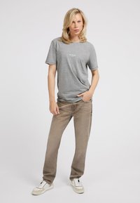 Grå kortærmet t-shirt med en hvid "GUESS" logo, parret med lysebrune lige ben jeans og hvide sneakers med beige accenter.