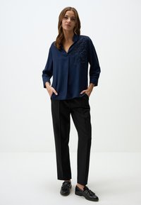 Blusa di seta blu marina con scollo a V e ricami floreali sulla spalla, abbinata a pantaloni neri a gamba dritta e scarpe nere.