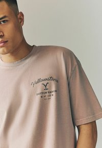 Lyserød bomulds T-shirt med rund halsudskæring, korte ærmer og et grafisk print med "Yellowstone Dutton Ranch" i mørk skrift.