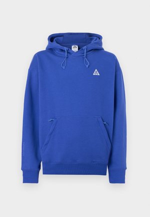 Blauwe hoodie gemaakt van zachte stof met een voorzak, capuchon met trekkoord en een klein driehoekig logo op de borst. Tekst op de mouw luidt "THE REMA FIT."