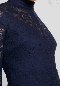 Robe en dentelle bleu marine avec un col haut, des motifs floraux complexes et un tissu texturé avec des sections transparentes sur les manches.