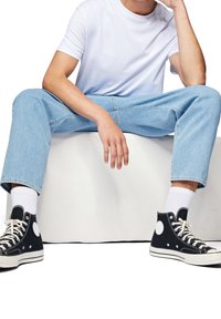 Personne portant un jean bleu clair, un t-shirt blanc, des chaussettes blanches et des baskets montantes noires, assise les jambes écartées sur un bloc blanc.