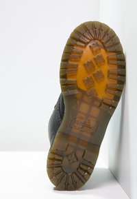 Semelle de bottes noires avec une bande de roulement translucide marron présentant des motifs géométriques et un texte en bloc, positionnée contre un fond blanc d'angle.