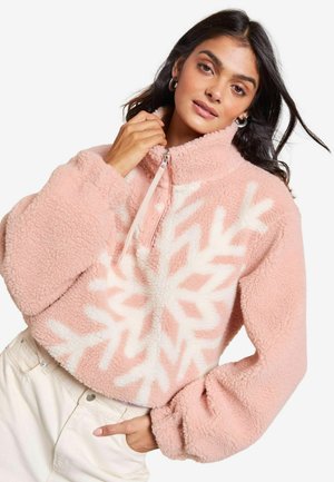 Pull en polaire rose pâle avec un motif de flocon de neige blanc, col montant et fermeture éclair. Comprend des manches larges et froncées ainsi qu'une coupe cropped.