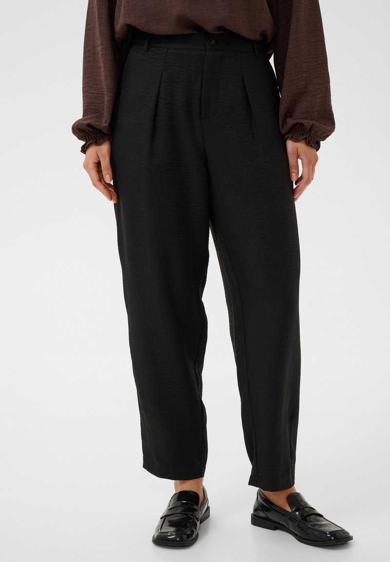 Personne portant un pantalon noir taille haute fuselé avec des plis à l'avant, associé à un haut marron à manches longues et des mocassins noirs brillants.