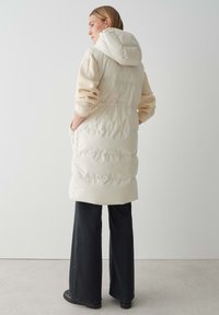 Puffige, cremefarbene Weste mit Kapuze, mit einem gesteppten Design und seitlichen Taschen, kombiniert mit einem hellen Pullover und dunklen weit geschnittenen Hosen.