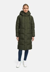 Lungo cappotto puffer verde oliva con cappuccio, caratterizzato da una chiusura zip, cordini elastici e un design trapuntato. Indossato sopra jeans e sneakers.