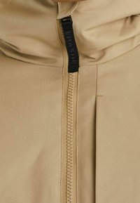 Beżowa kurtka z wysokim kołnierzem, z centralnym zamkiem błyskawicznym i tkaniną o fakturze. Zamek błyskawiczny posiada uchwyt u góry.