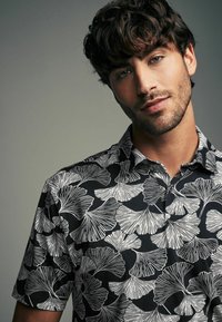Kortærmet polo-shirt med sort stof og hvide ginkgo-blad mønstre, krave og knapdetaljer. Letvægts tekstur.