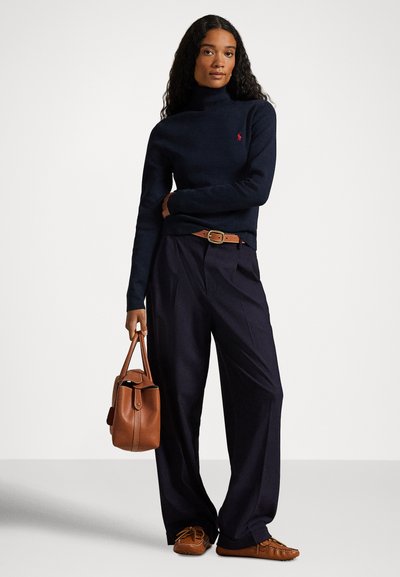 Ženska, oblečena v mornarsko moder pulover z visokim ovratnikom, široke mornarsko modre hlače, rjav pas in mokasine, ob strani drži rjavo usnjeno torbico.