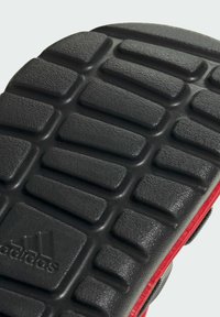 Semelle en caoutchouc texturé noir avec des blocs de tread profonds et un bord rouge partiel, présentant le logo Adidas en relief en bas.