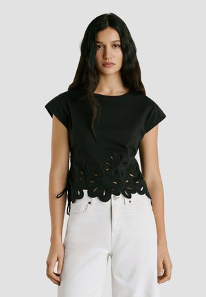 Vrouw draagt een zwart topje met korte mouwen, een bloemenuitgesneden zoom en witte, hoog taille jeans, staand tegen een effen grijze achtergrond.