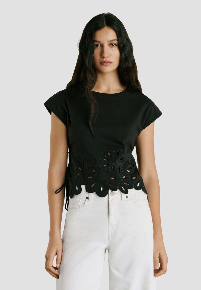 Vrouw draagt een zwart topje met korte mouwen, een bloemenuitgesneden zoom en witte, hoog taille jeans, staand tegen een effen grijze achtergrond.