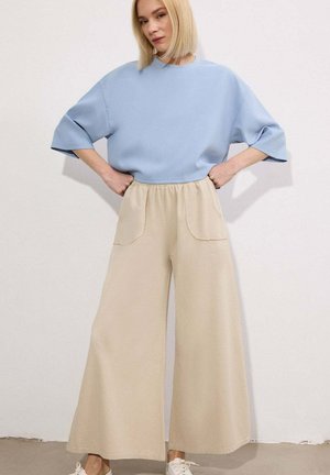 Kobieta o blond bob fryzurze, ubrana w jasnoniebieską, dużą bluzkę z krótkim rękawem i szerokie beżowe spodnie z przednimi kieszeniami, stojąca z rękami na biodrach.