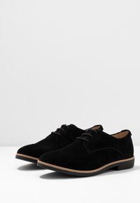Chaussures noires en suède à lacets avec un bout arrondi et une semelle en caoutchouc marron clair contrastante. Texture lisse avec des détails de couture minimalistes.