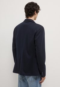 Mann mit lockigem Haar, der einen dunkelblauen Blazer und hellblaue Jeans trägt und mit dem Rücken zu einem schlichten weißen Hintergrund steht.