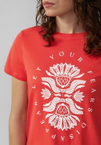T-shirt en coton rouge avec un grand motif floral blanc et le texte "LAISSEZ VOS PEURS DISPARAÎTRE" centré sur le devant. Manches courtes, col rond.