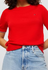 Kobieta ubrana w jasnoczerwoną, krótką bluzkę z dzianiny, włożoną w jasnoniebieskie, wysokie dżinsy, stojąca z rękami za plecami.