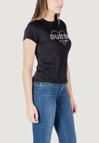 Zwarte katoenen T-shirt met korte mouwen, voorzien van een zilveren glitter logo-ontwerp met een hartmotief. Gecombineerd met blauwe spijkerbroek.