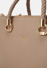 Sac à main beige avec une surface texturée, des poignées tressées, des accessoires en or, et le nom de marque embossé "LIU-JO". Fermeture à glissière incluse.