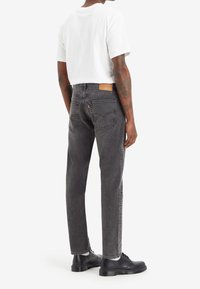 Vaqueros de denim negros con un corte recto, que presentan un parche de cuero en la cinturilla y un detalle de costura sutil. Llevados con una camiseta blanca y zapatos negros.