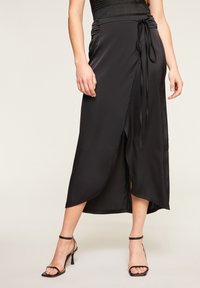 Černá saténová zavinovací sukně s vysokým rozparkem, nastavitelným zavazováním v pase a hladkým povrchem, doplněná černými páskovými podpatky.