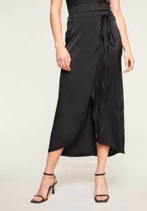 Jupe enveloppante en satin noir avec une fente haute, lien ajustable à la taille et texture lisse, associée à des sandales à talons noires à lanières.