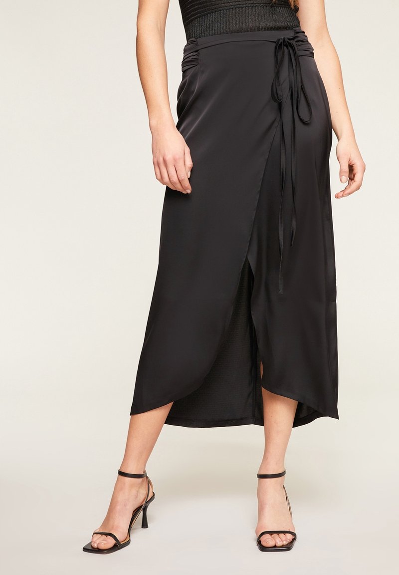Černá saténová zavinovací sukně s vysokým rozparkem, nastavitelným zavazováním v pase a hladkým povrchem, doplněná černými páskovými podpatky.