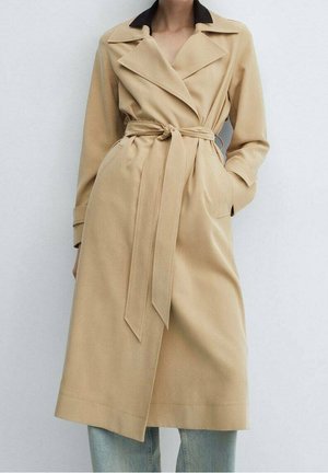 Kvinde iført en beige trenchcoat med bælte og brede slag over lyseblå jeans, stående mod en ensfarvet lys baggrund.