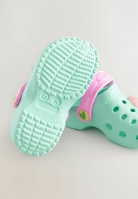 Zuecos verde claro con suela de goma texturizada, que cuentan con orificios de ventilación, una correa trasera rosa y pequeños acentos de corazón en la correa.