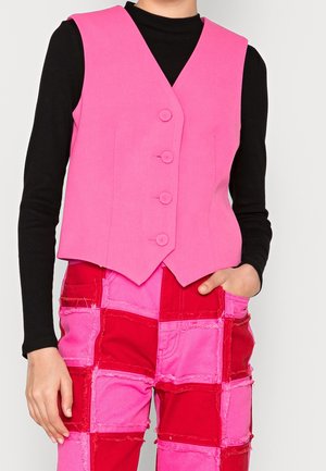 Person wearing bright pink buttoned vest over black long-sleeve shirt and patchwork pants with red and pink square patches.