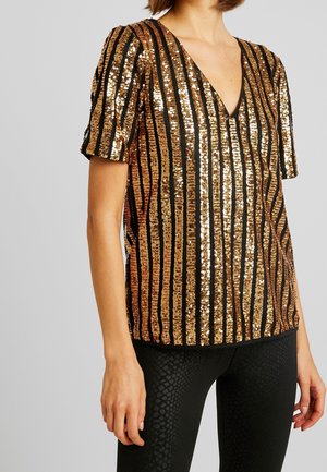 Vrouw met een gouden en zwarte top met korte mouwen en verticale paillettenstrepen, gecombineerd met zwarte, gestructureerde leggings tegen een effen achtergrond.