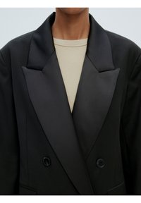 Zwarte dubbel opgesneden blazer met een satijnen uitstraling, brede revers en twee zwarte knopen, gecombineerd met een textuurbeige top.