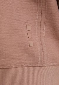 Gros plan sur un tissu marron clair avec des coutures visibles et trois petits patchs carrés en caoutchouc alignés verticalement.