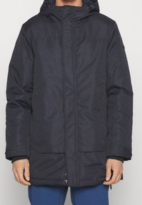 Giacca nera isolata con zip frontale, cappuccio regolabile e ampie tasche anteriori. Realizzata in un materiale liscio e resistente.