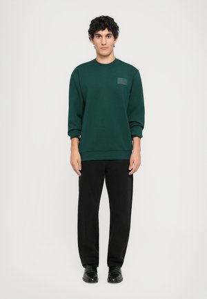 Jonge man die staat in een donkergroen sweatshirt, zwarte broek en zwarte schoenen tegen een eenvoudige witte achtergrond.