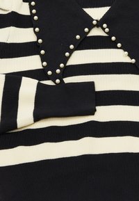 Sweter w czarno-kremowe paski o prążkowanej fakturze, z wyraźnym kołnierzem ozdobionym perłowymi akcentami wzdłuż dekoltu.