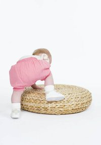 Un bébé en short rose grimpe sur un tapis rond en paille. Le tapis a une texture tissée, et le bébé porte des chaussures blanches ainsi qu'une chemise avec un nœud.