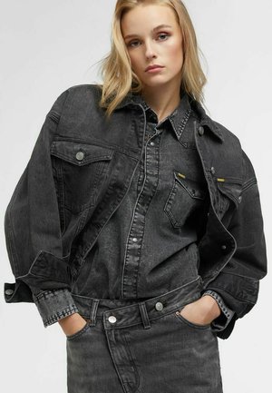 Blonde Frau mit den Händen in den Taschen, trägt eine aufeinander abgestimmte dunkelgraue Jeansjacke, ein Hemd und eine Jeans vor einem einfarbigen grauen Hintergrund.