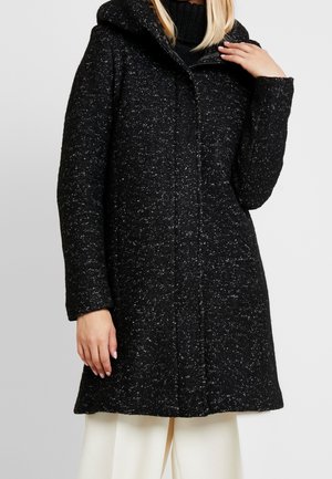 Vrouw draagt een zwarte woljas met vlekken en capuchon over een zwarte coltrui, gecombineerd met een witte broek.