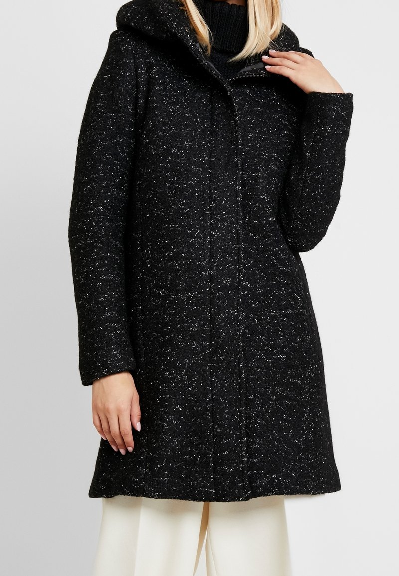 Femme portant un manteau en laine noir moucheté avec une capuche, sur un pull à col roulé noir, associé à un pantalon blanc.